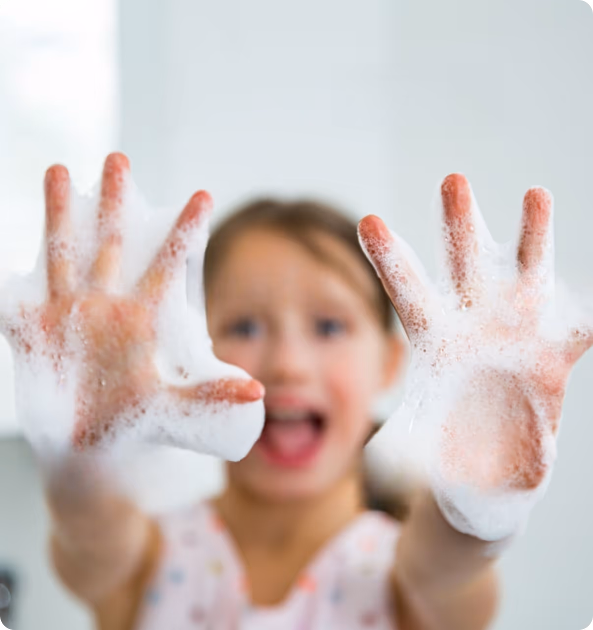 Kind, das in einem Kita Waschraum glücklich lacht und seine beiden eingeschäumten Handflächen in die Kamera hält-