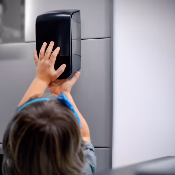 Kind in einem Kita-Waschraum, dass einen dunklen Seifenspender nutzt, um sich die Hände zu waschen